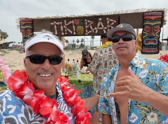 Two men in hawaiian shirts and hats at a tiki bar.